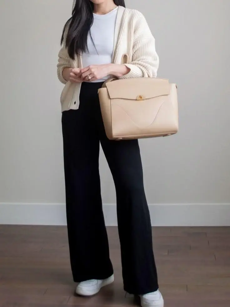 Woman wearing a travel outfit with a white T-shirt, black trousers, a lightweight cardigan, white sneakers, and a carry-on suitcase as part of a capsule wardrobe.